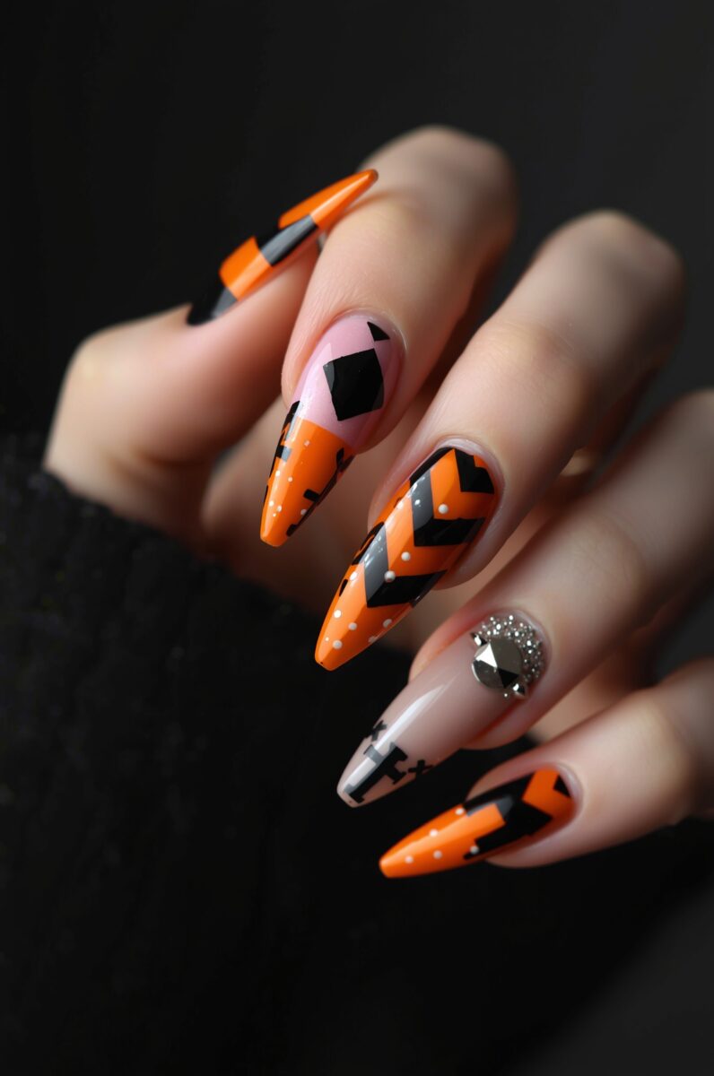 Hand with long stiletto nails featuring orange and black geometric Halloween nail art with chevron patterns and rhinestone accent
