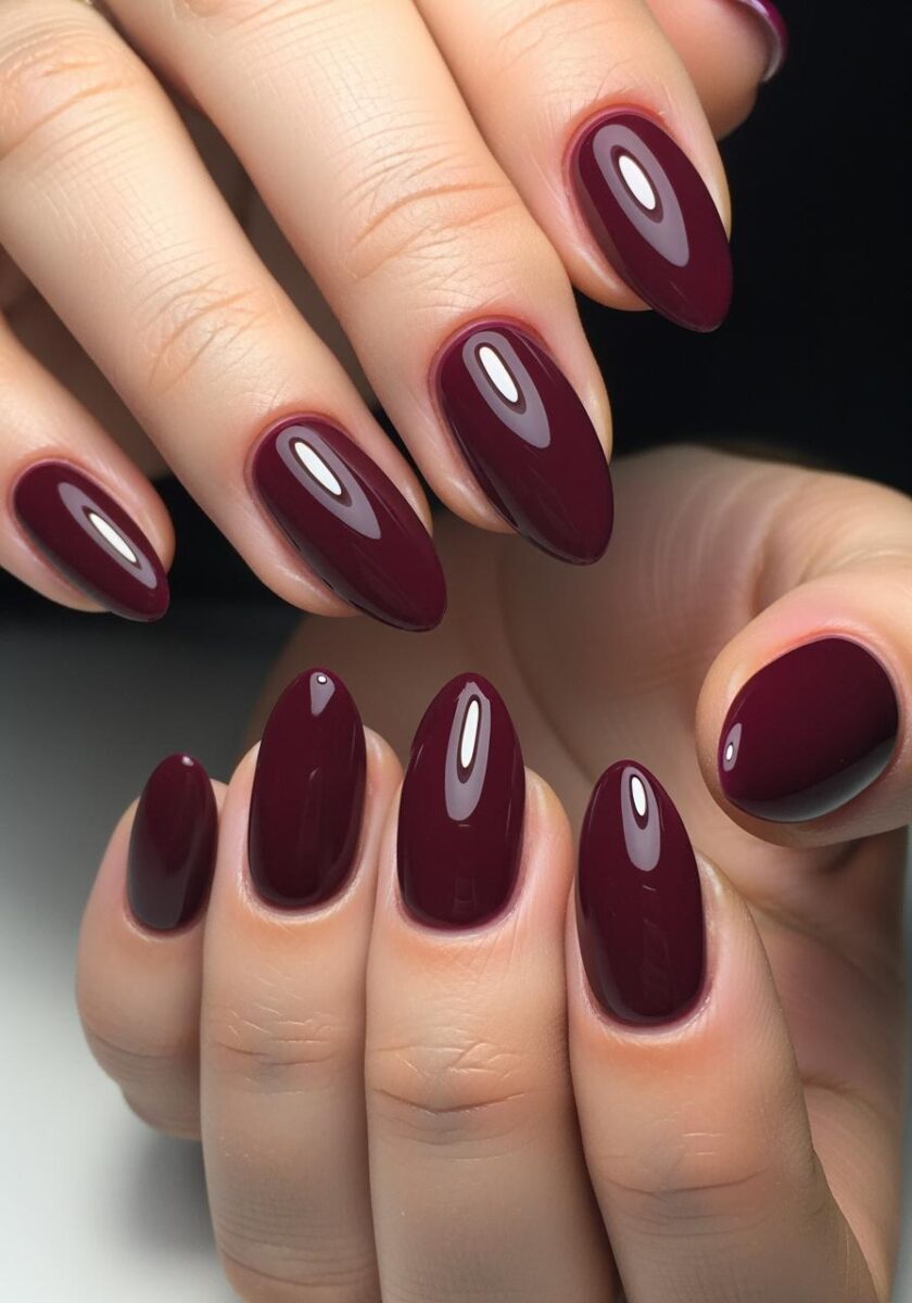 Two hands with glossy deep burgundy almond-shaped gel manicure nails against dark background