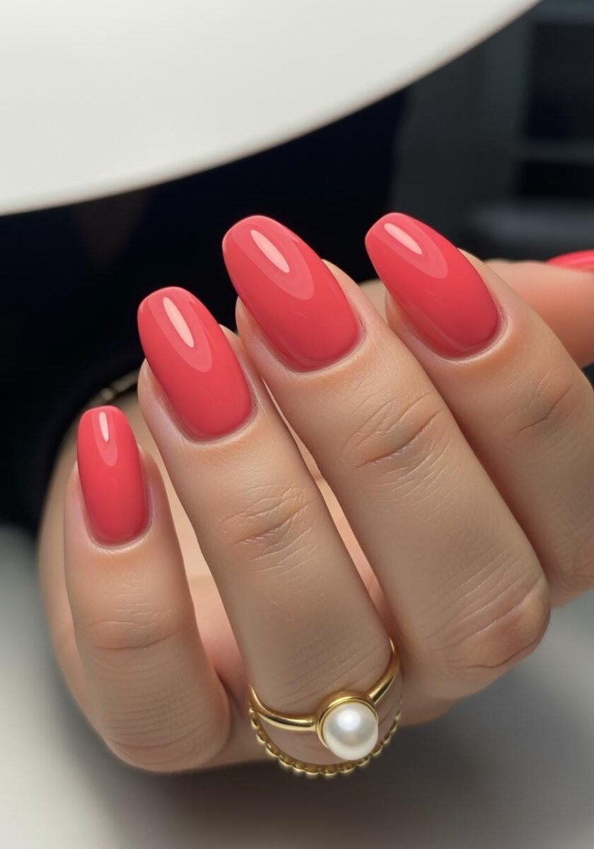 Close-up of a hand with glossy coral-pink oval nails wearing a gold pearl ring