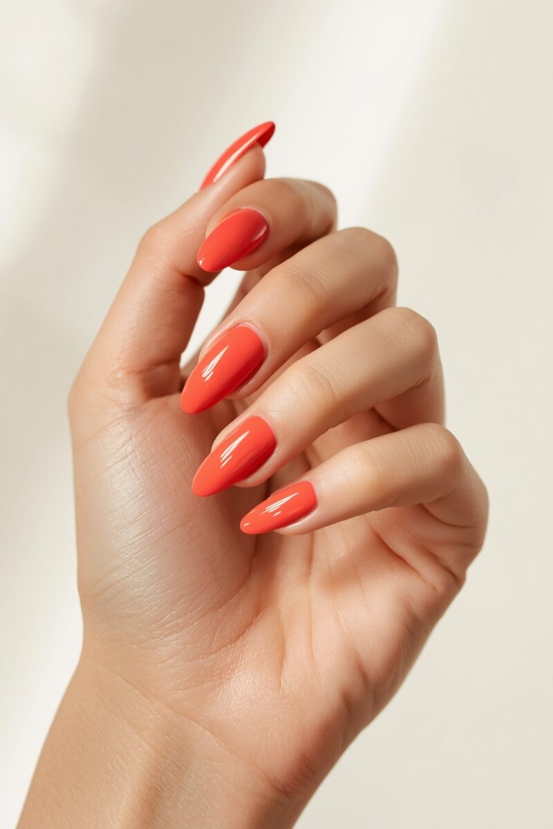 Close-up of a hand with glossy coral-orange almond-shaped nail polish on medium-length nails