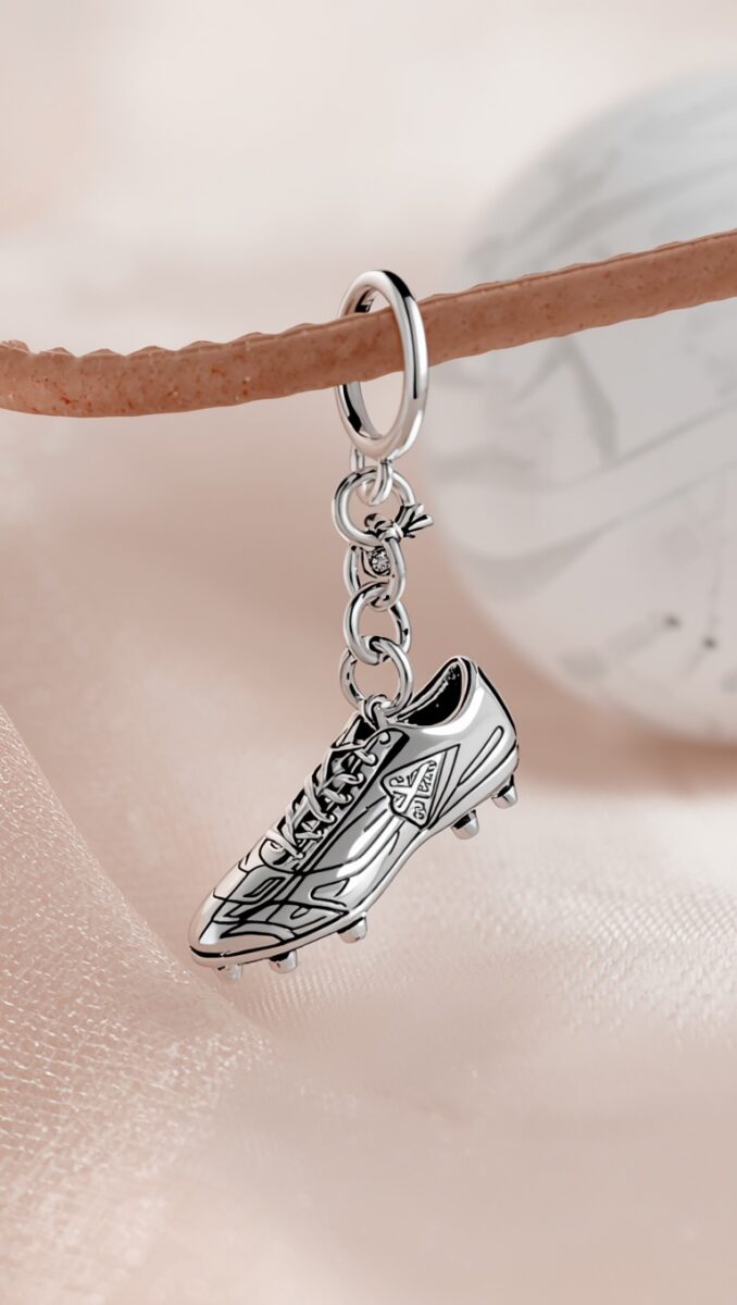 Silver soccer cleat charm pendant hanging from a chain and ring on a leather strap, soft pink background