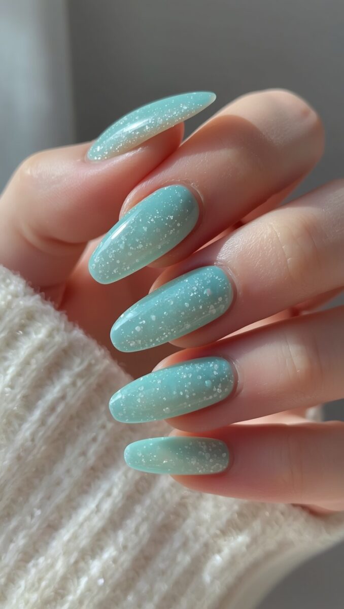 Close-up of almond-shaped nails painted in mint green with white glitter sparkle finish on a hand resting on white knit sweater