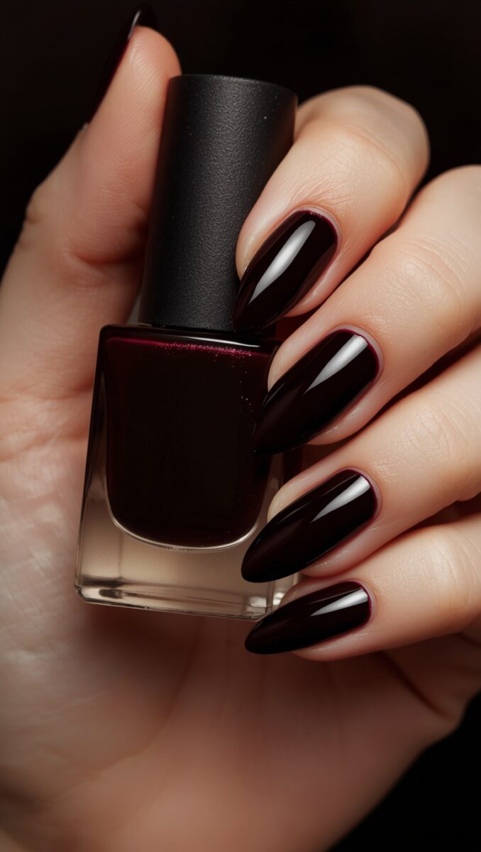 Hand with long stiletto nails painted in deep dark burgundy polish holding a matching nail polish bottle