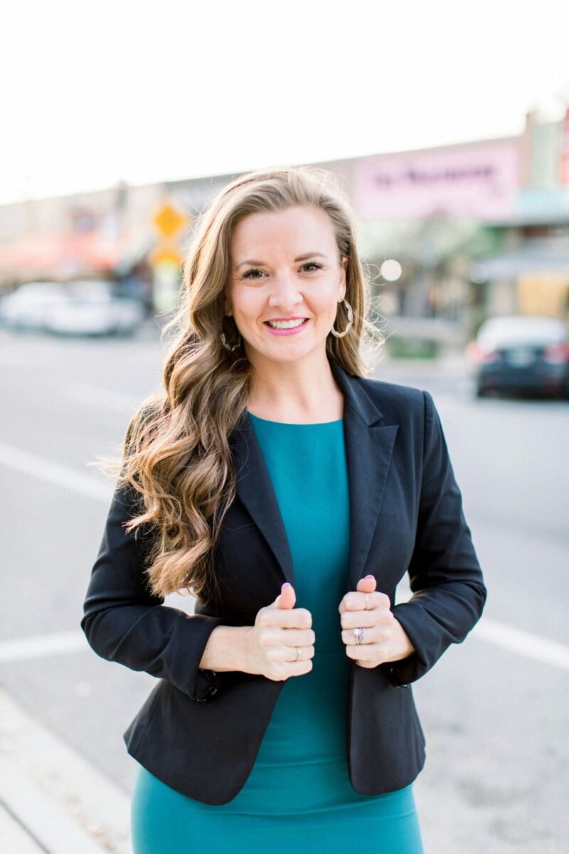 Smiling woman holding the lapels of a fitted navy blazer over a teal sheath dress
