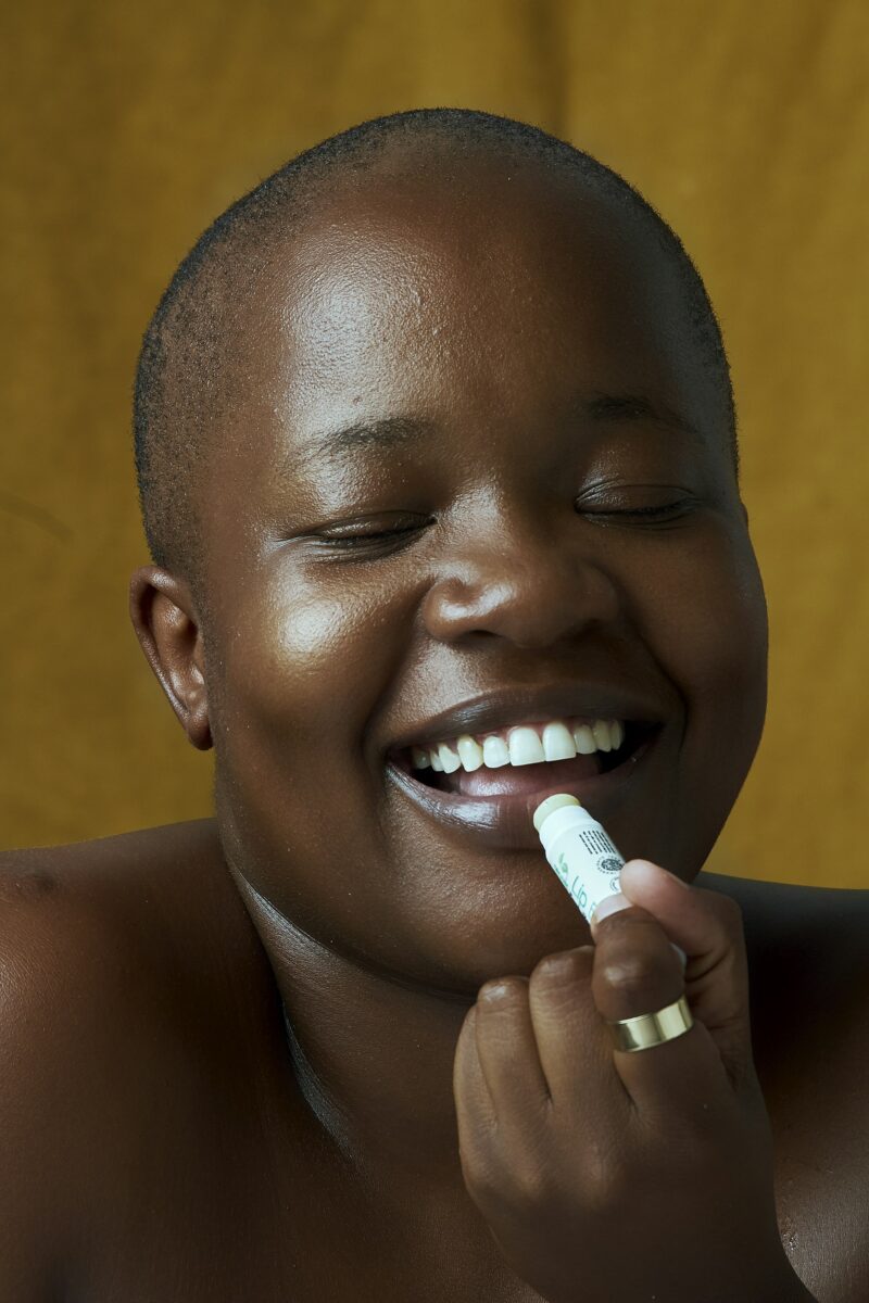 A smiling woman with a shaved head putting on lip balm with her eyes closed