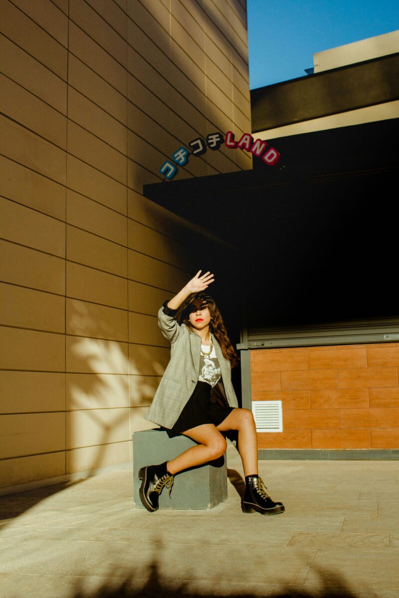Woman in a gray check blazer, graphic T-shirt, short black skirt and lace-up boots sitting on a concrete block