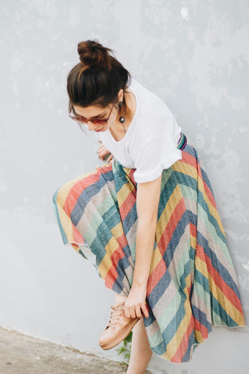 A rainbow-striped pleated midi skirt with a plain white tee, sunglasses and tan slip-on shoes