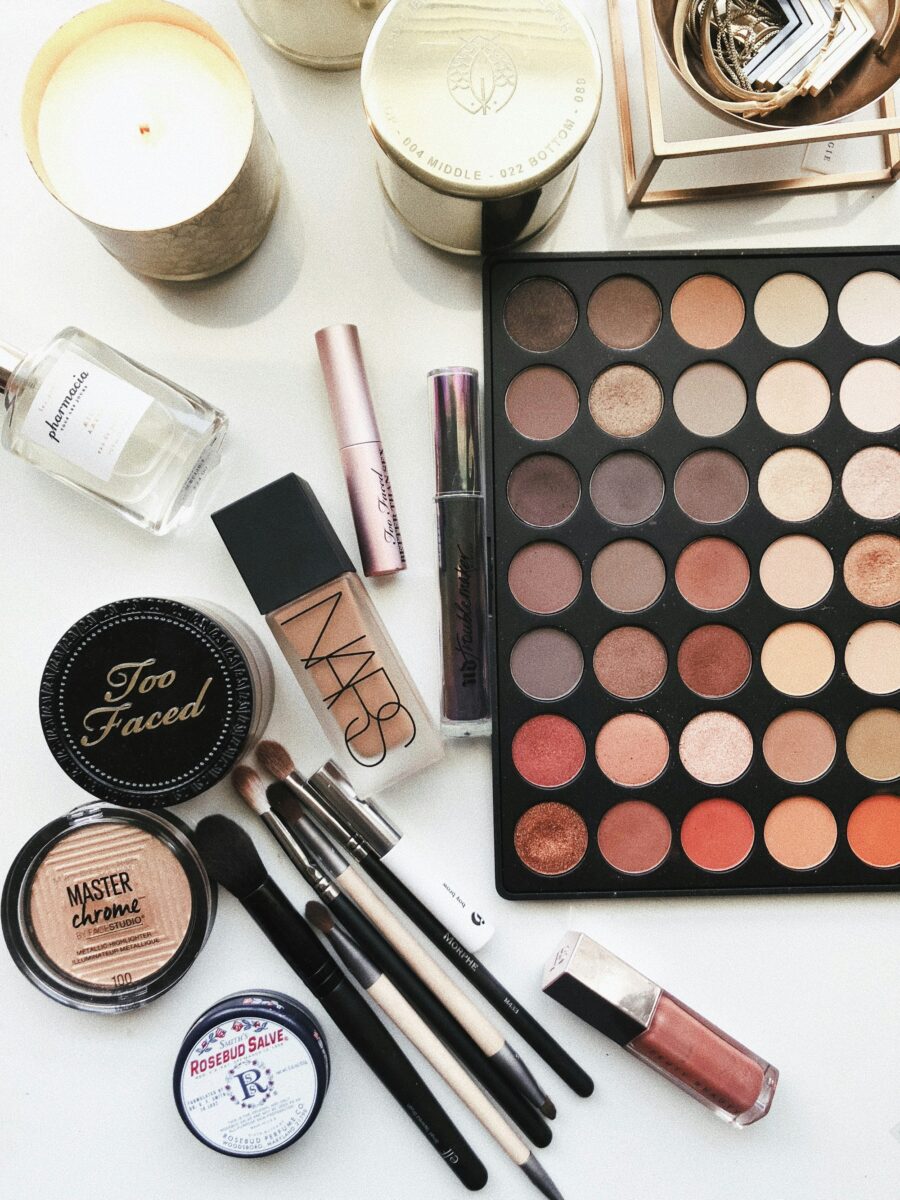 Makeup laid out on a white table: an eyeshadow palette, foundation, mascara, brushes, highlighter and lip gloss