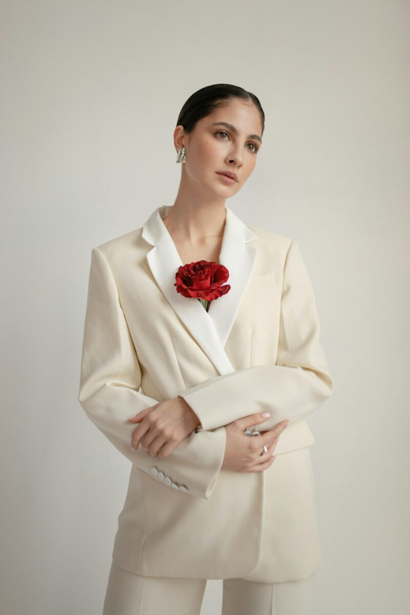 Woman in a closed cream suit jacket with white satin lapels and a red rose at the neckline