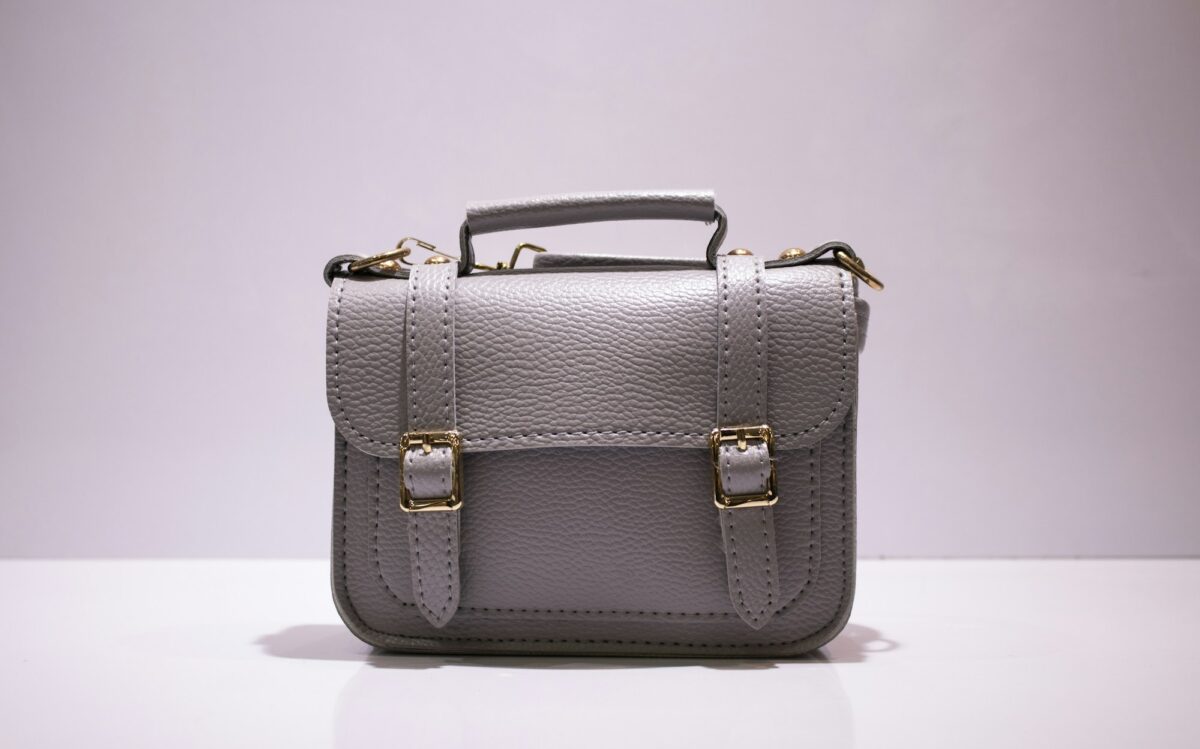 A small gray leather satchel with gold buckles, a top handle and rings for a shoulder strap