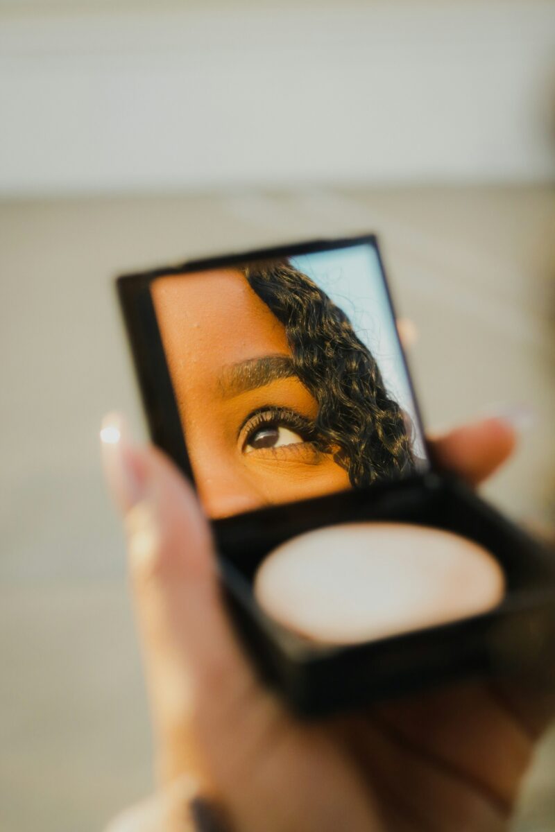 A hand holding up an open powder compact whose mirror reflects a woman's eye