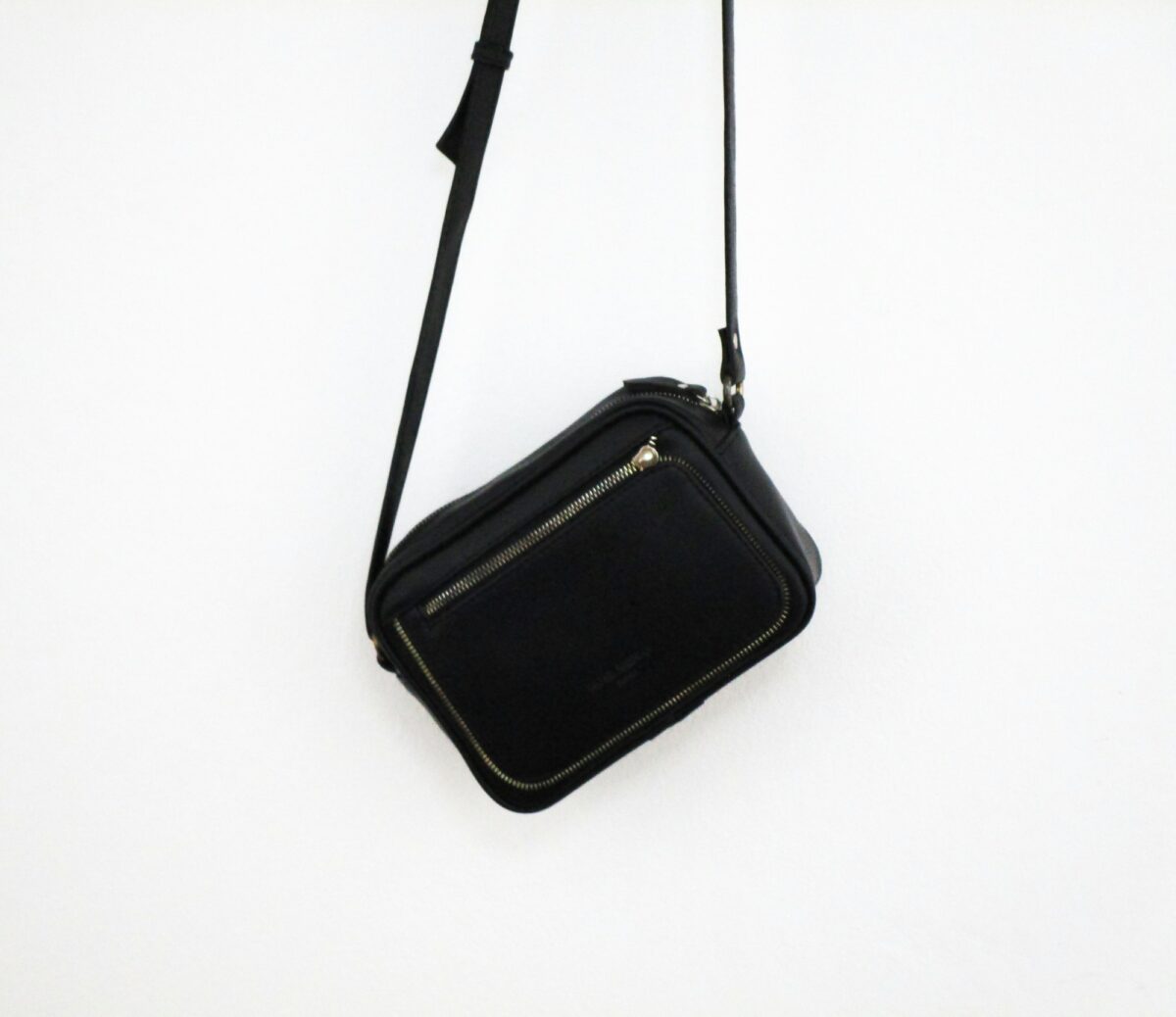 A plain black leather crossbody bag with a zip top and a front zip pocket, hanging by its strap