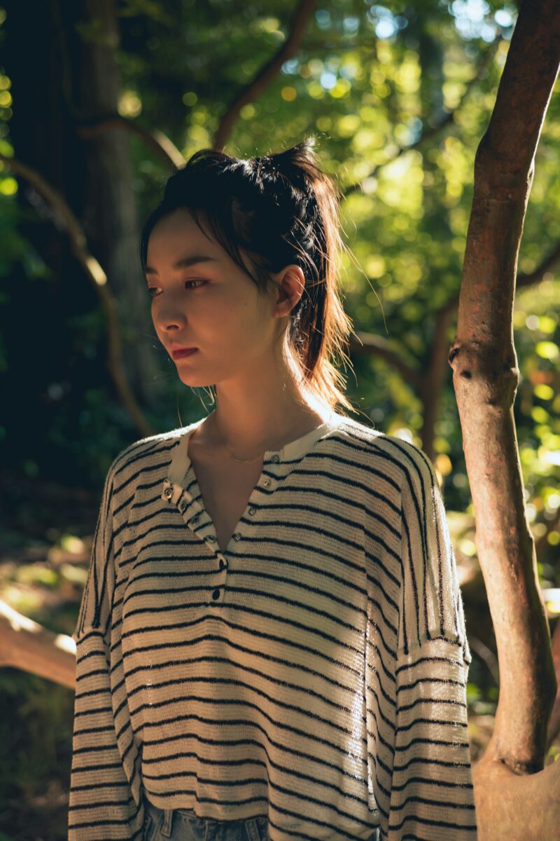 An oversized cream henley with black stripes and a buttoned placket, worn loose in late-afternoon sun under the trees