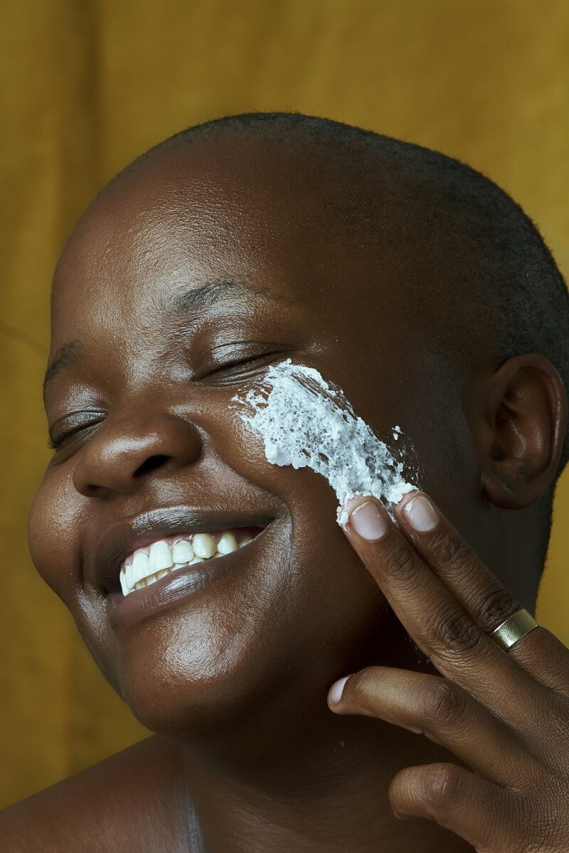 A smiling woman with her eyes closed smoothing a white face product onto her cheek with her fingertips