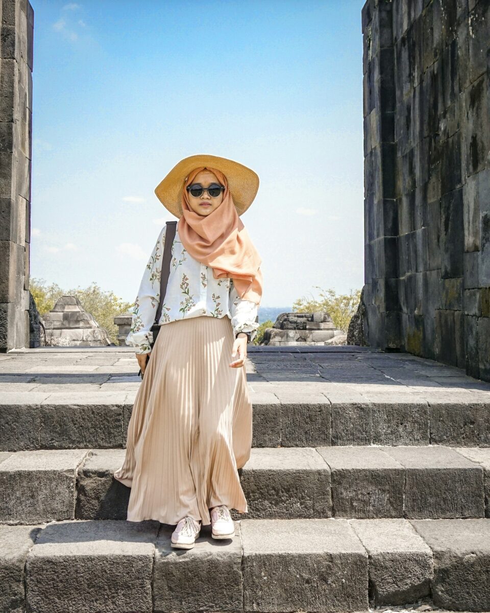 A beige pleated maxi-length skirt with a floral blouse, a peach headscarf, a straw hat and white sneakers on old stone steps
