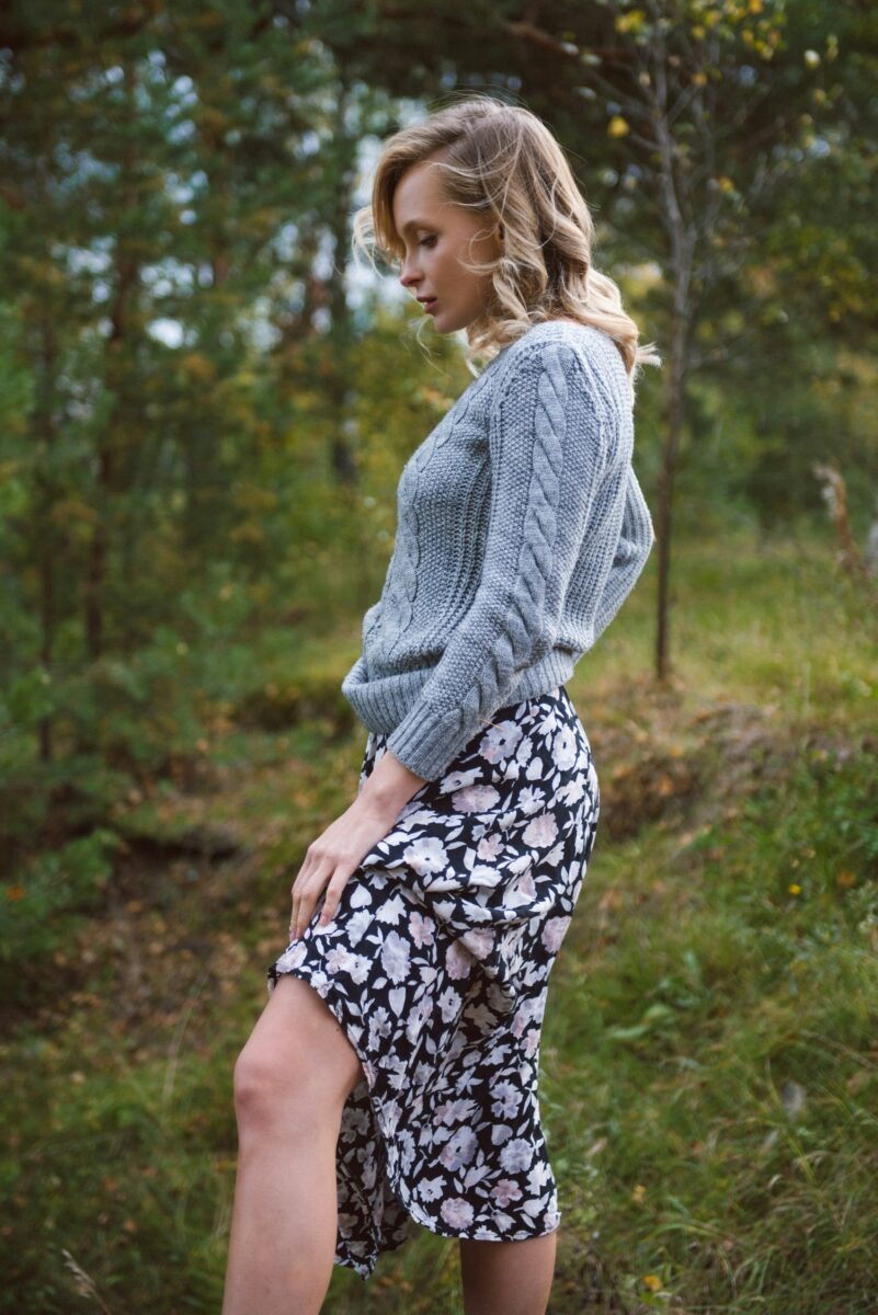 A gray cable-knit sweater ending at the waist over a black and pink floral slip midi skirt, in a forest in early fall
