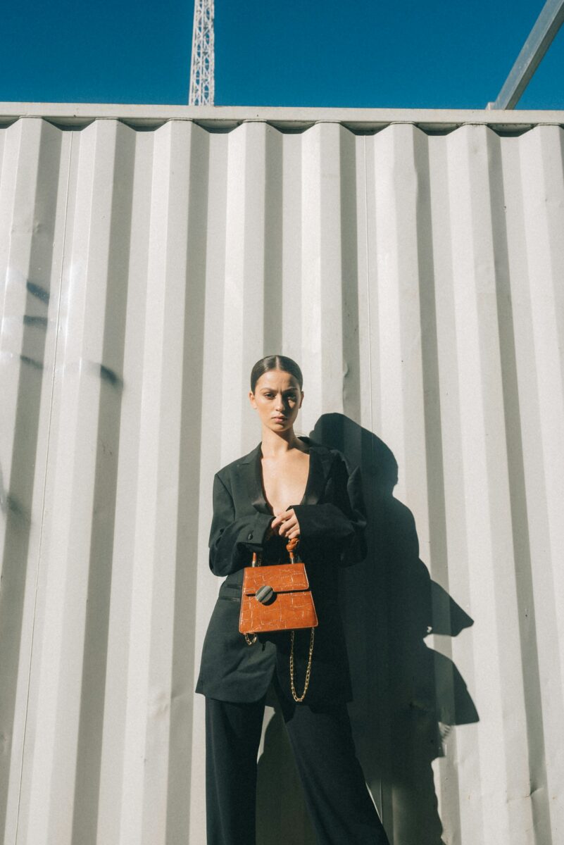 A woman in a black suit holding a structured orange top-handle bag with a chain strap