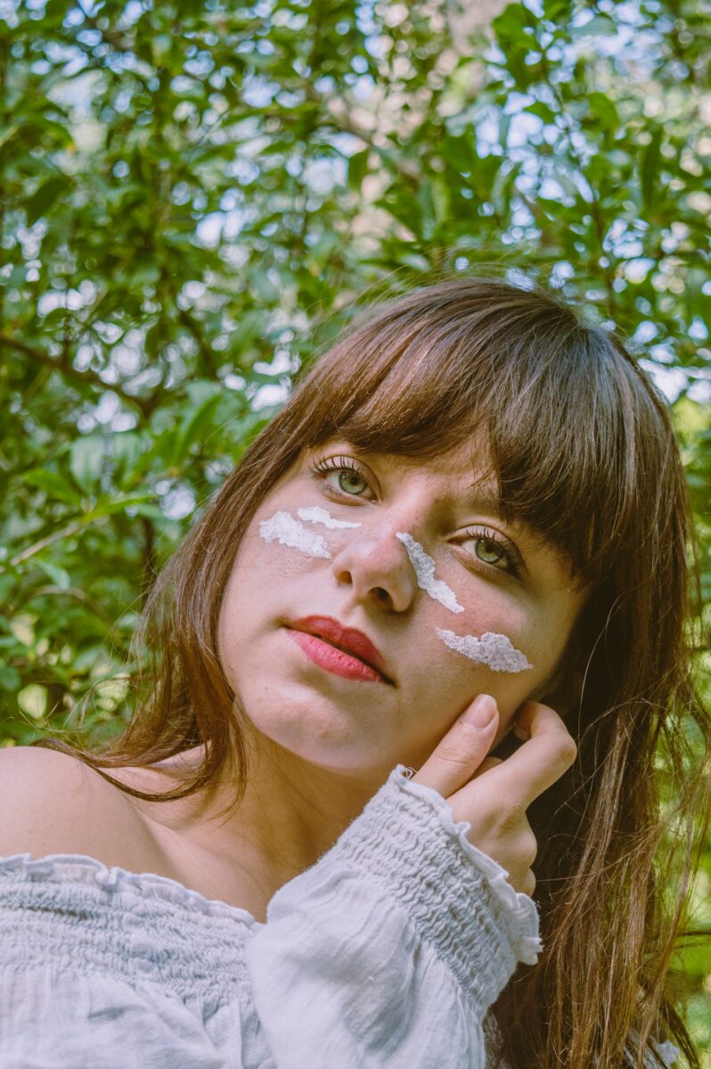 A woman with bangs and dabs of white sunscreen on her cheeks and nose, outdoors in front of green leaves