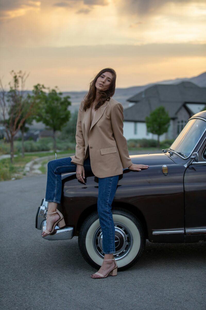 Woman in an oversized camel blazer, straight blue jeans and block-heel sandals leaning on a vintage car