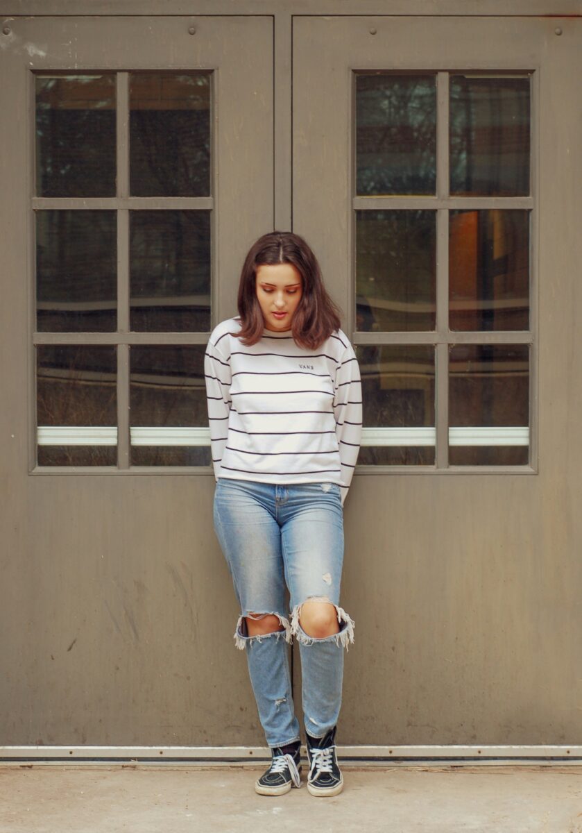 A white long-sleeve top with thin black stripes, ripped light-wash jeans and black high-top sneakers, leaning on a pair of paneled doors