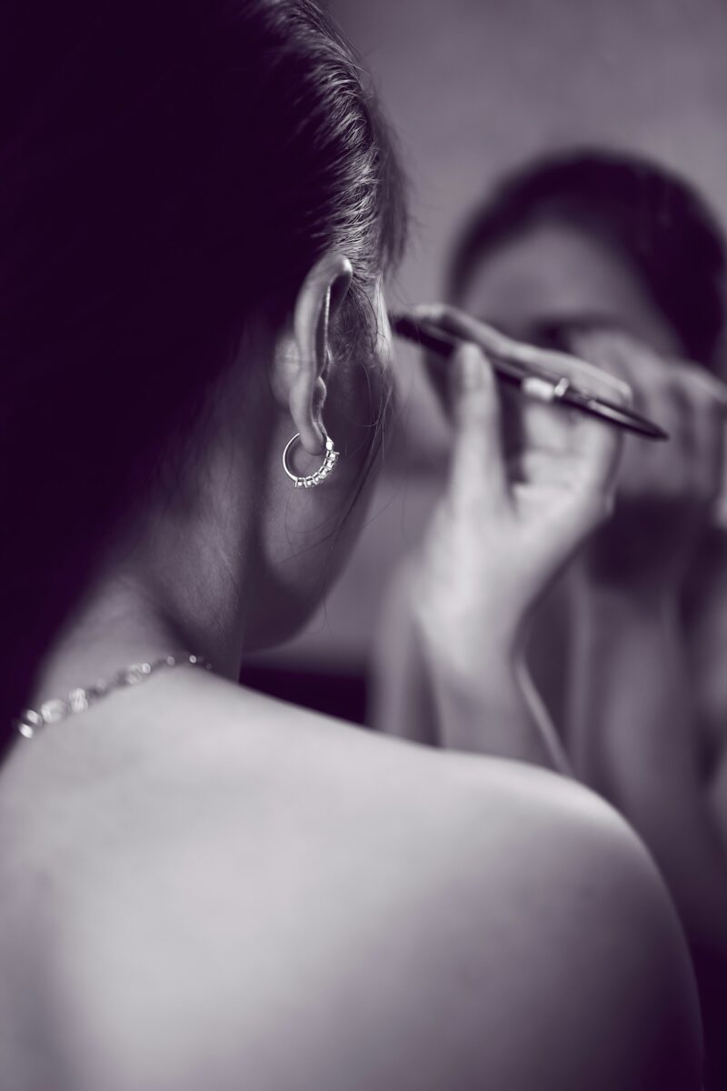 A woman filling in her eyebrow with a pencil in front of a mirror, in purple-toned black and white