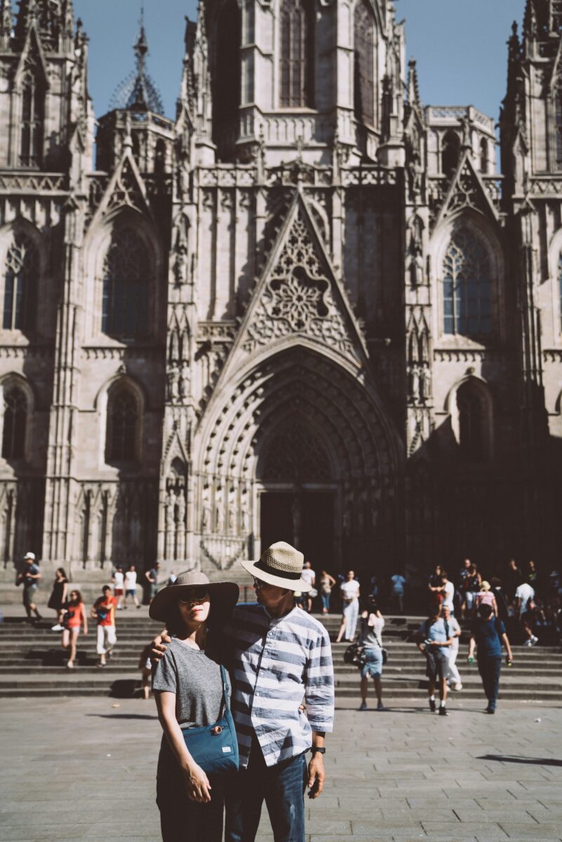 A man in a blue and white striped button-down and jeans with his arm around a woman in a gray tee, in front of a Gothic cathedral