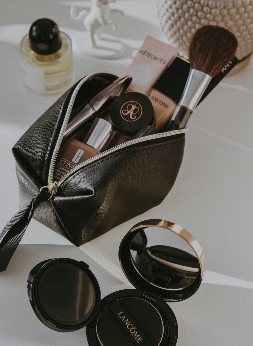 A small black makeup bag unzipped with brushes, foundation and a cushion compact, two open compacts beside it