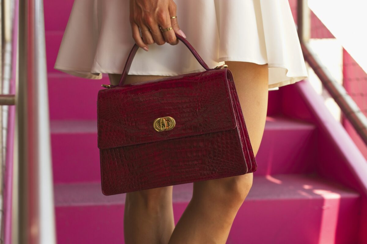 A burgundy croc-effect leather bag with a gold clasp, held by its top handle on pink stairs