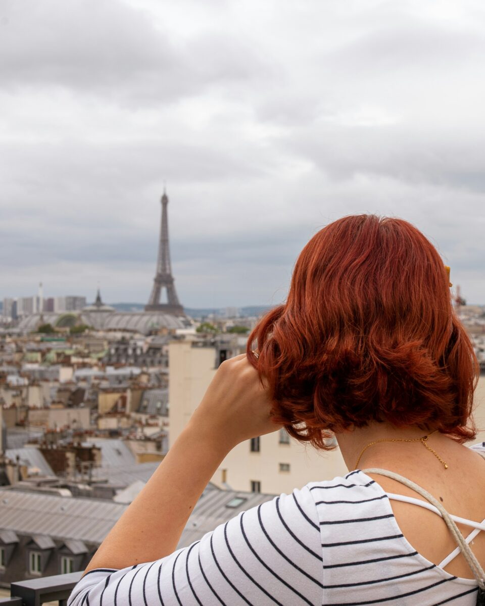 A woman in a white top with thin navy stripes and a laced open back, looking over Paris rooftops toward the Eiffel Tower