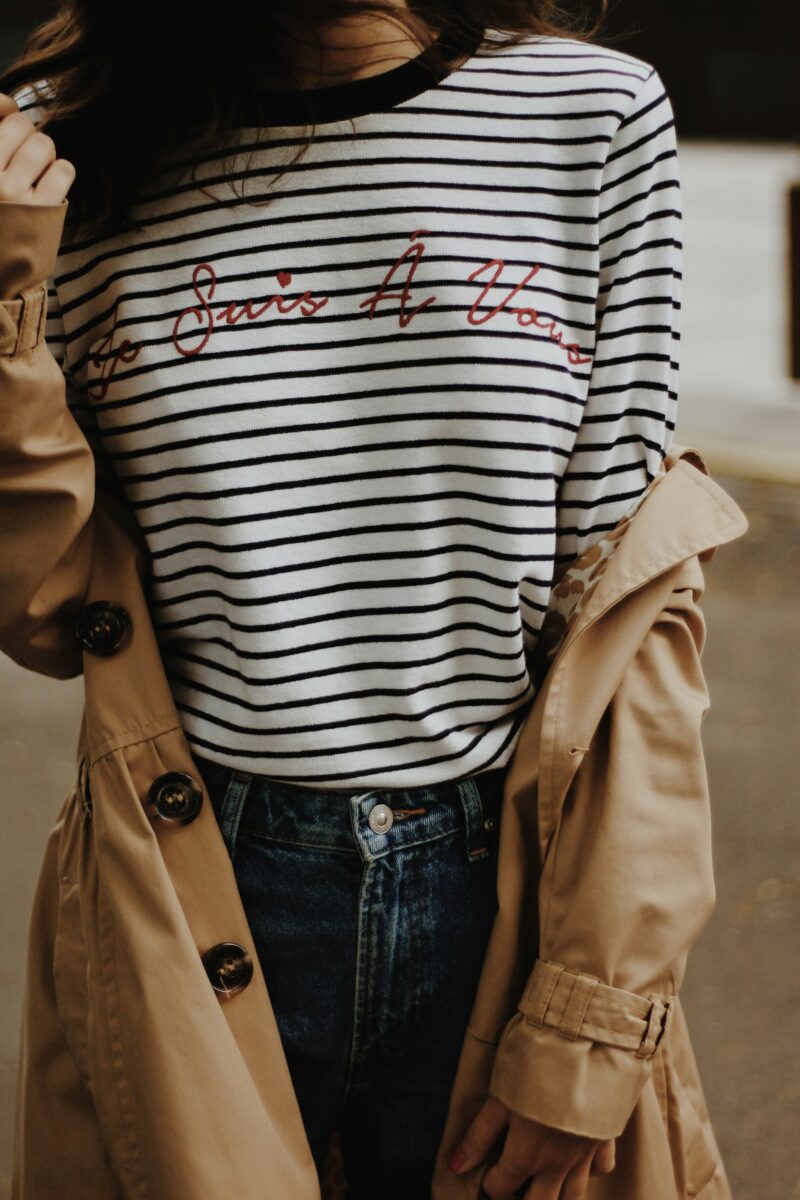 Striped shirt outfit for fall: a white long-sleeve top with thin navy stripes, high-waisted dark jeans and a camel trench coat slipping off one shoulder
