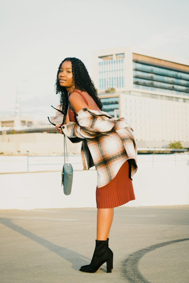 A rust pleated knit midi skirt with a plaid shirt jacket and black heeled ankle boots, a band of bare leg between the hem and the boot