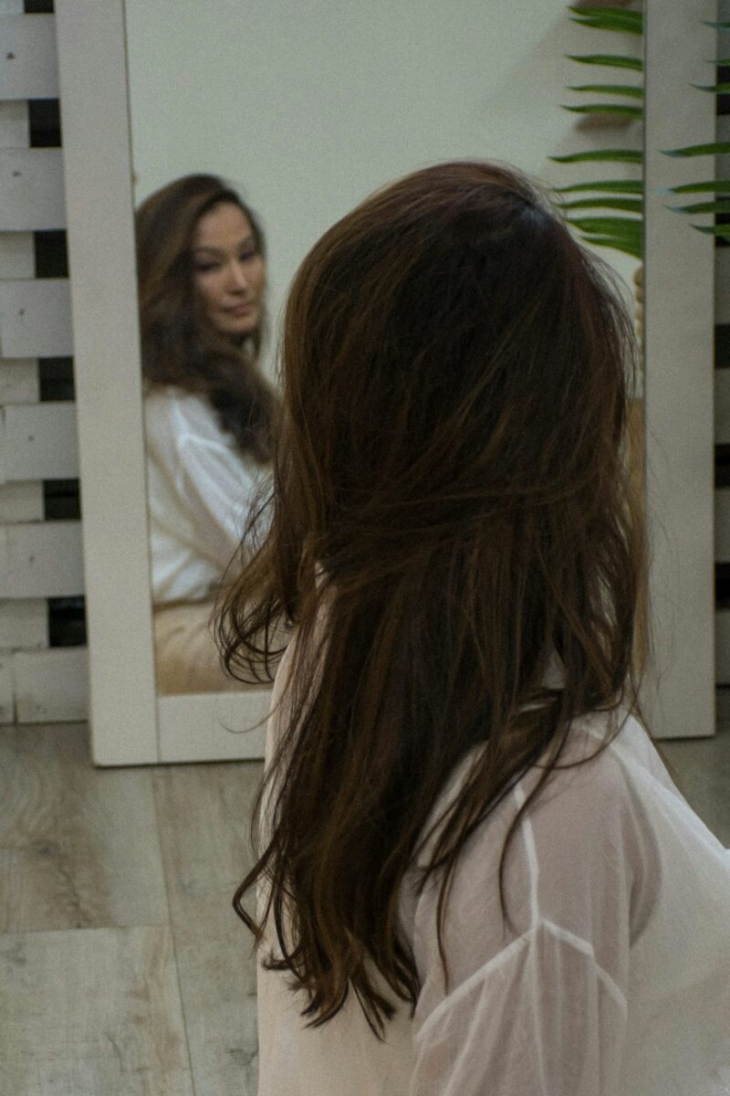 A woman with long brown hair looking closely at her face in a white-framed mirror