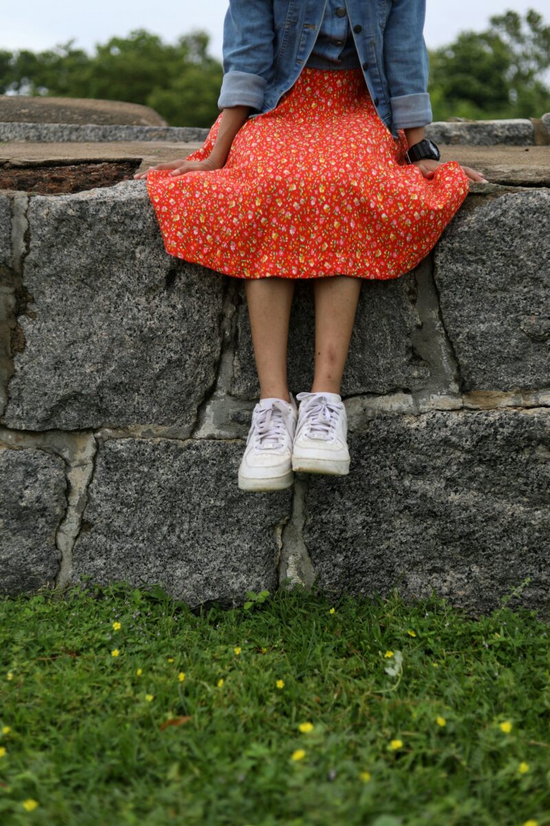 A red floral midi skirt with a cropped denim jacket and white sneakers, sitting on a granite wall