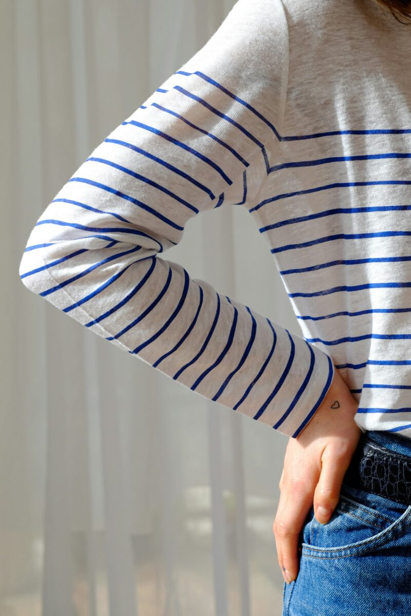 A close-up of a white top with thin cobalt stripes half-tucked into dark jeans with a black belt, one hand in the front pocket