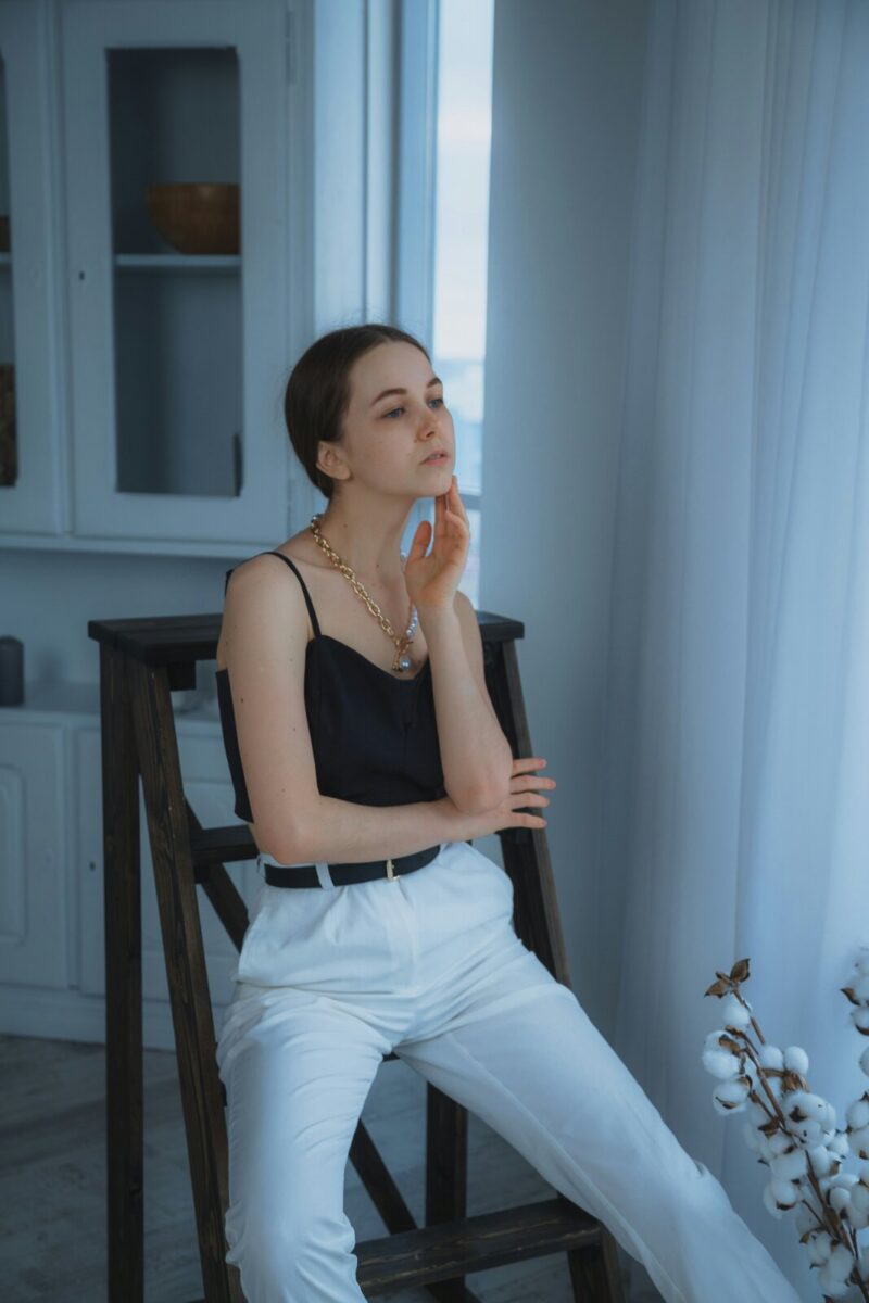 A black camisole, a gold chain necklace and belted white trousers, on a tall wooden chair by a window