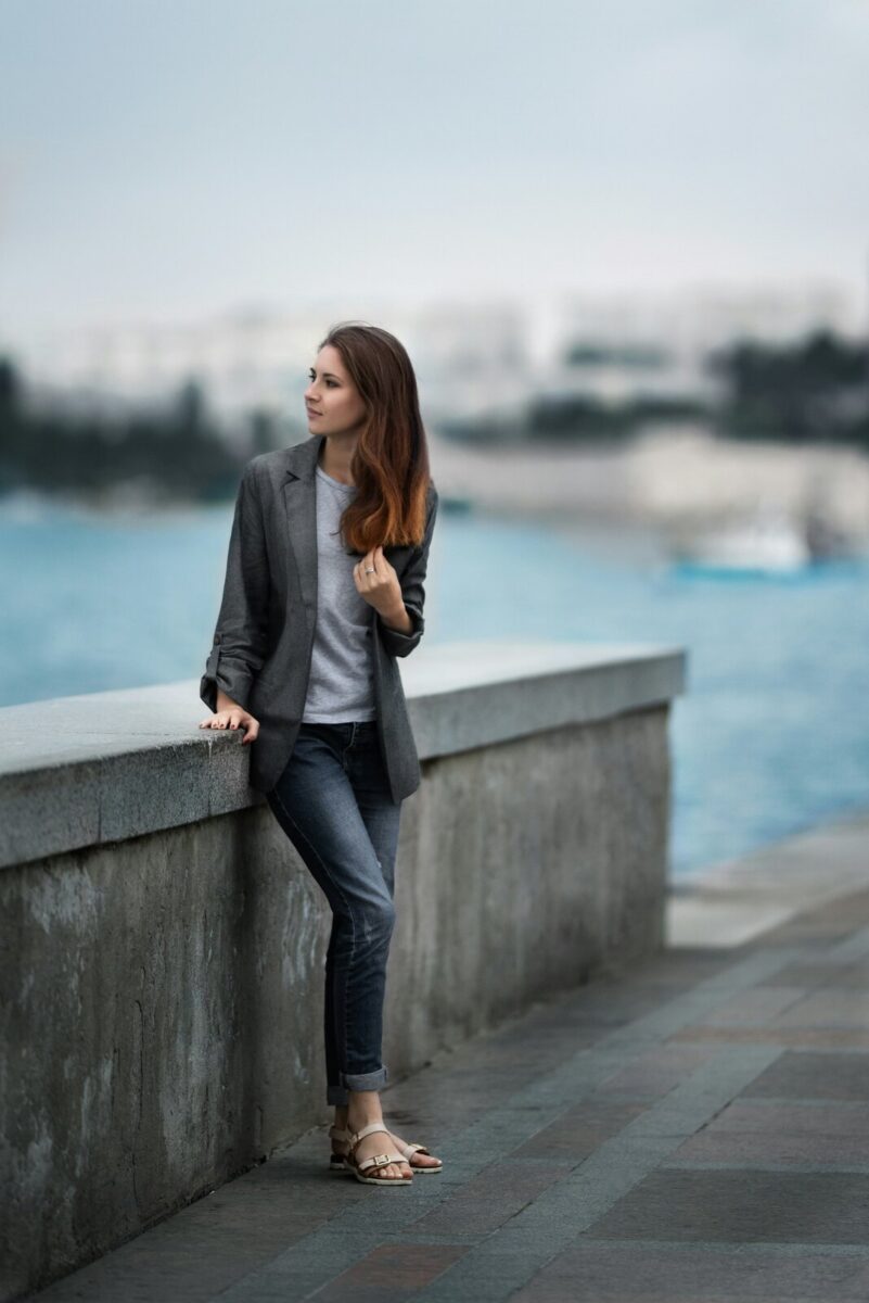 A gray blazer and gray tee with rolled blue jeans and flat sandals, by a seawall