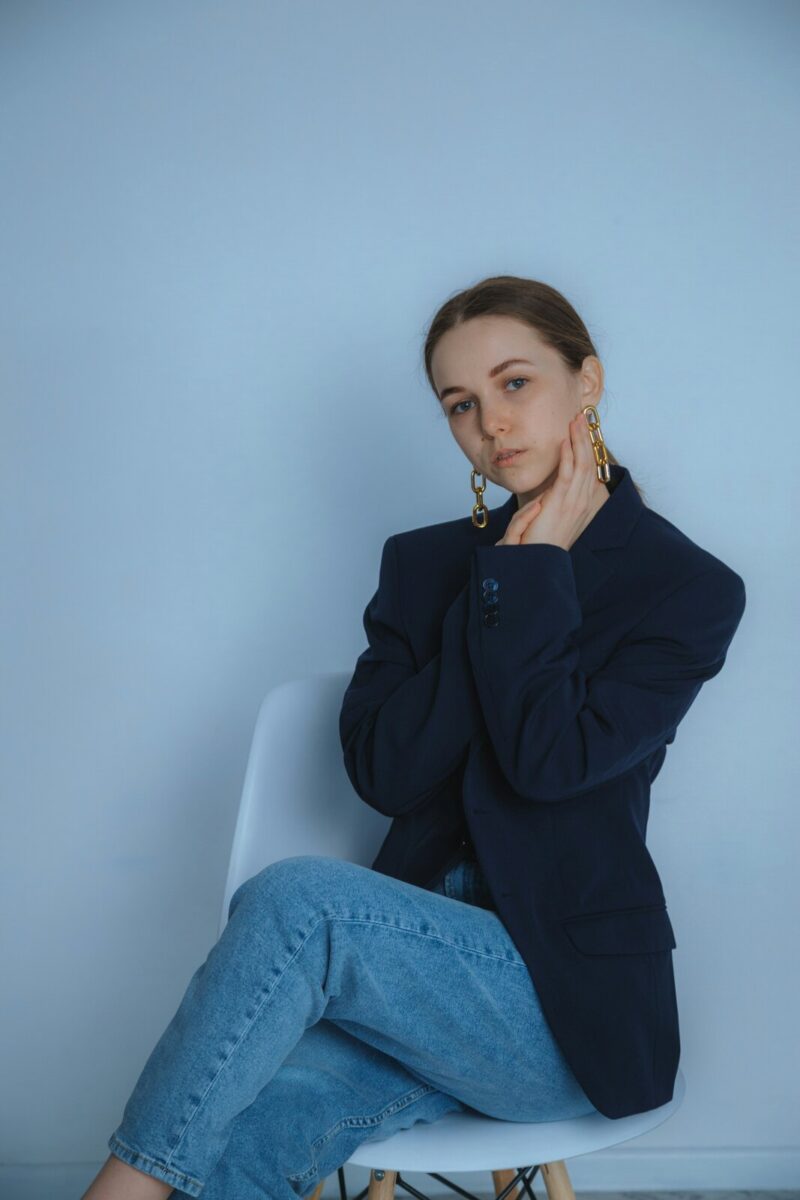 Long gold chain-link earrings with a navy blazer and light jeans, on a white chair against a pale blue wall