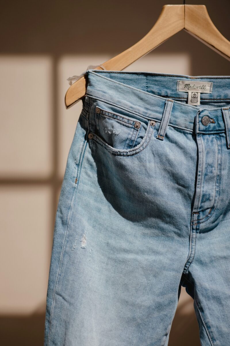 Light-wash jeans with a small rip hanging from a wooden hanger in window light