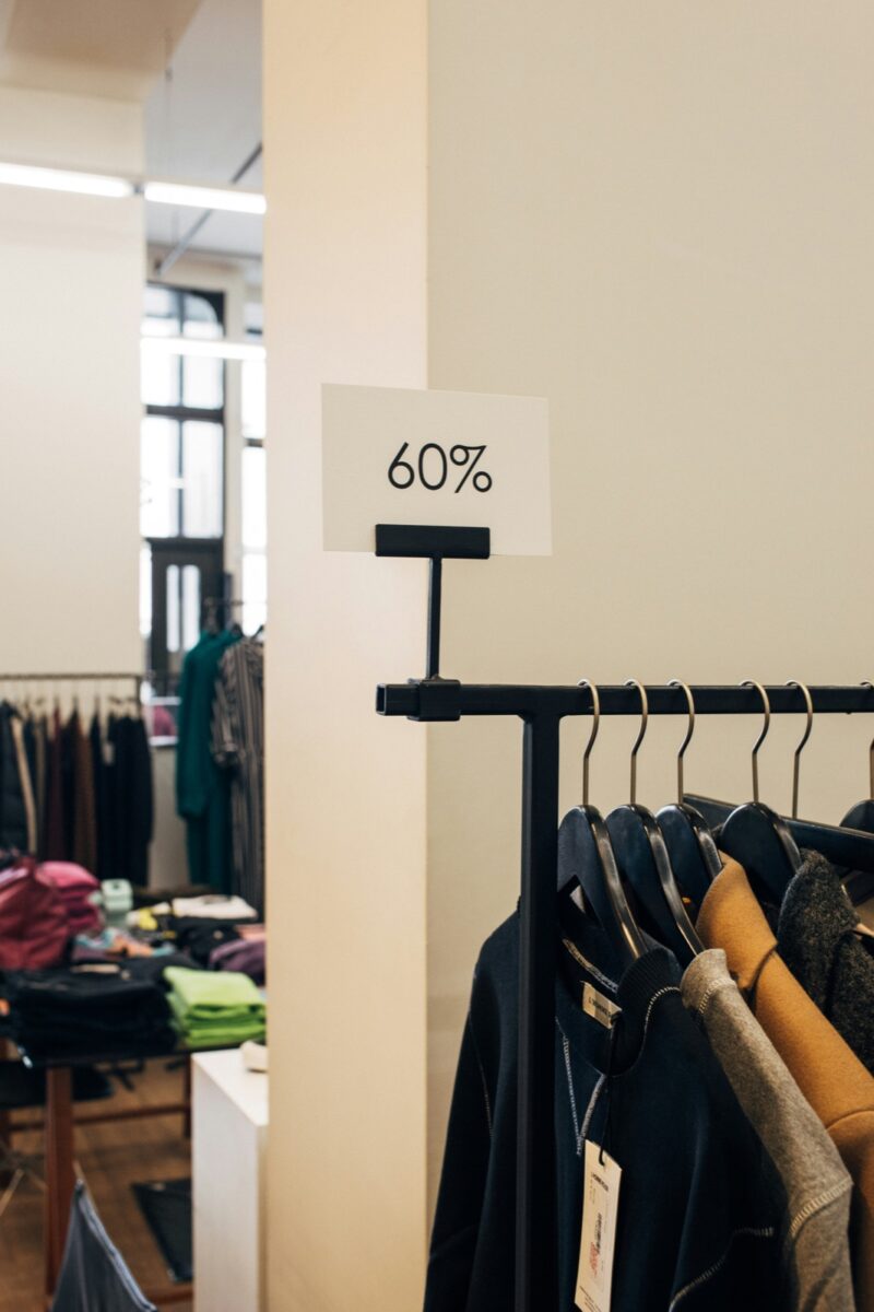 A white 60% sign on a rack of dark jackets and a camel coat in a clothing shop