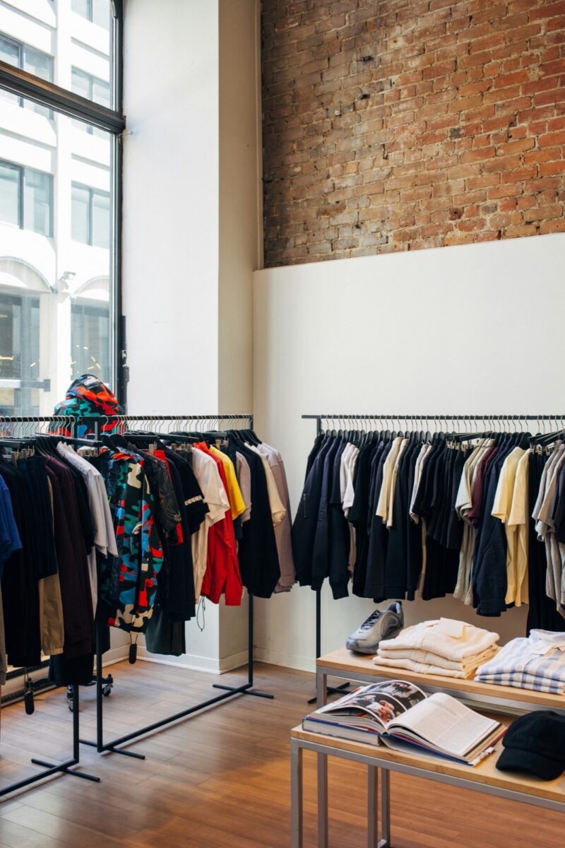 Two rails of shirts and jackets beside a table of folded shirts in a shop with a brick wall