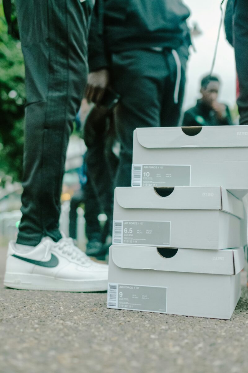 Three white sneaker boxes stacked on a street beside a pair of white low-top sneakers