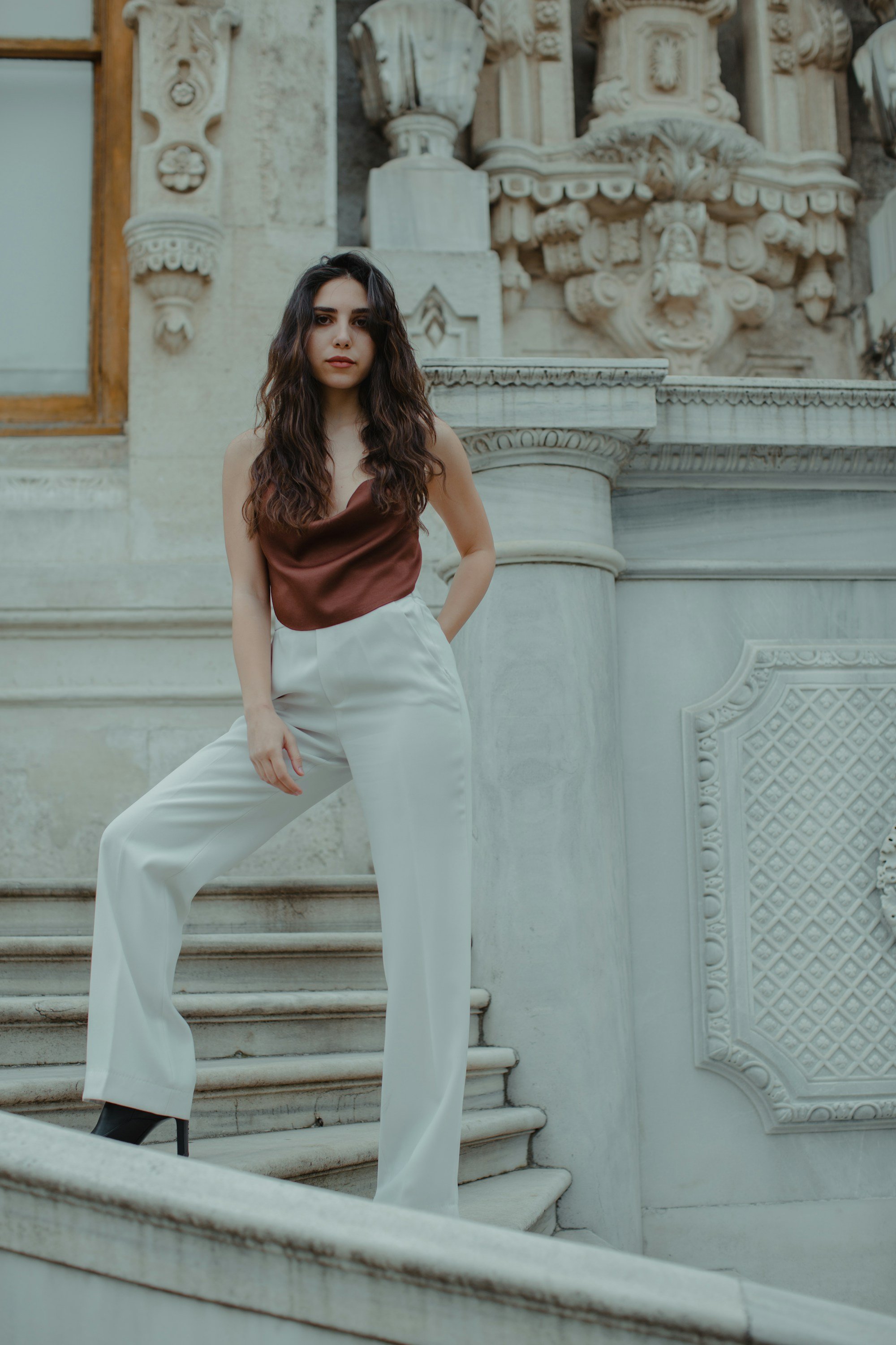 woman in a rust satin cowl neck cami tucked into ivory wide-leg trousers on stone stairs