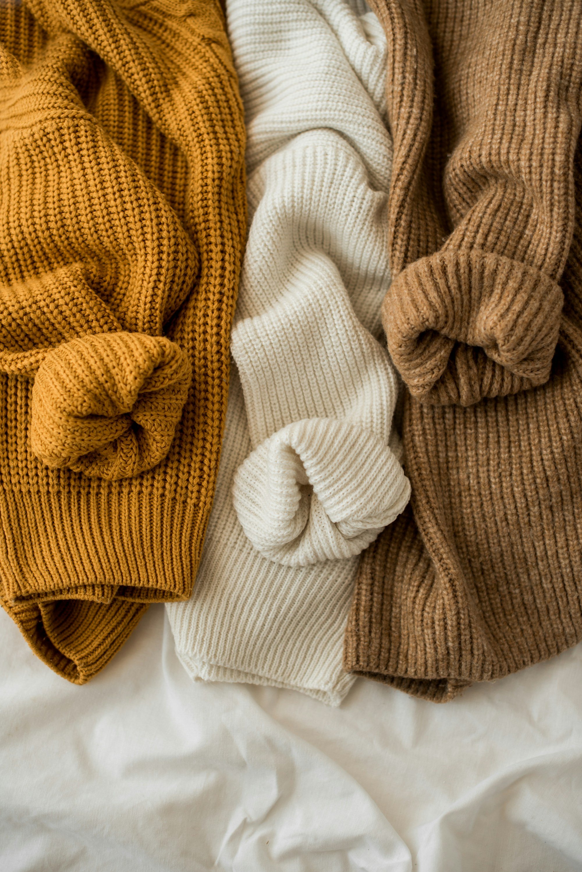 mustard, cream and camel rib knit sweaters laid side by side on a white bed